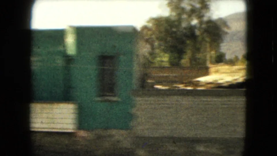 1962 Mexico-area home-movie view of a green-painted building with a window, boundary wall, trees, and a distant hillside.