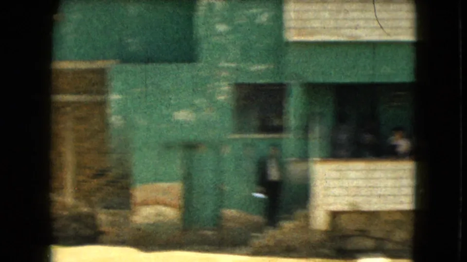 1962 Mexico-area frame showing a green-painted building, low walls, windows, dry ground, and one person in dark clothing near the exterior.
