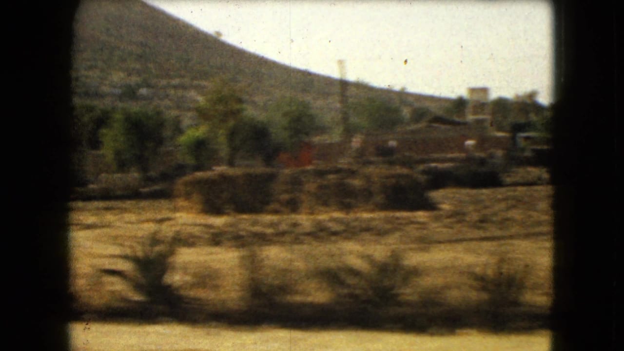 Dry field, shrubs, trees, and small buildings beneath a hillside.