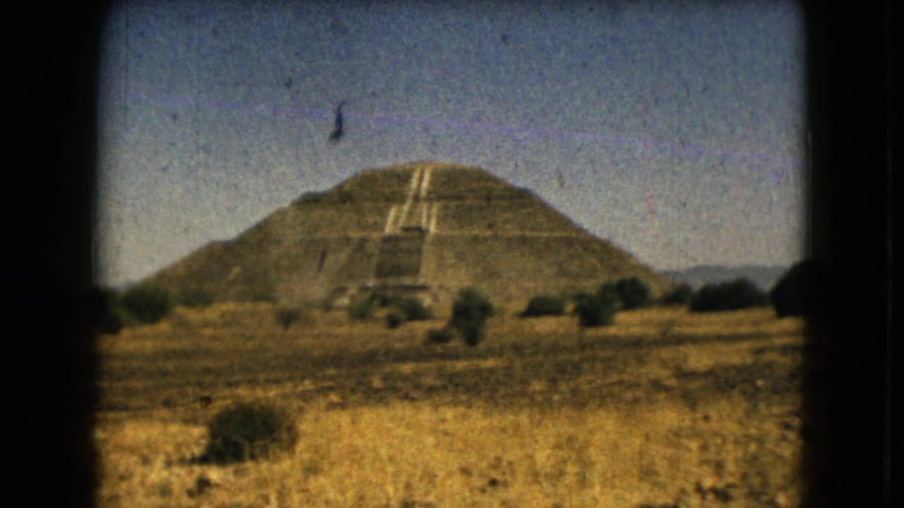 Stepped pyramid monument beyond a dry field under a clear sky.
