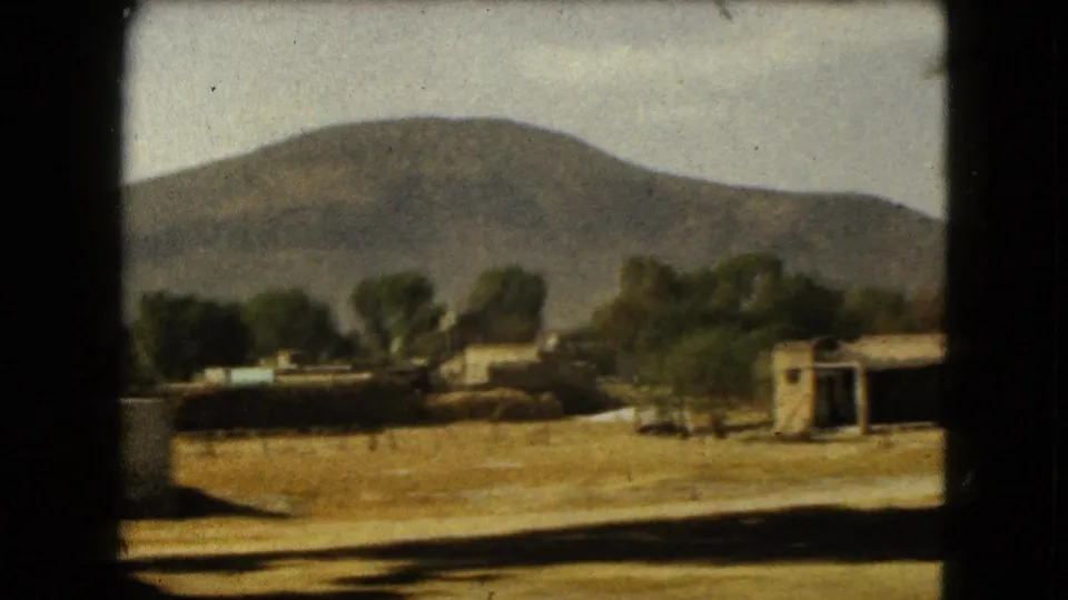 1962 Mexico City landscape frame with small rural structures, trees, dry ground, and a hazy mountain rising behind them.
