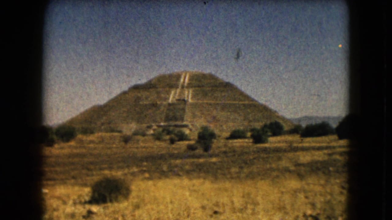 Large stepped pyramid across dry grass with shrubs and a clear sky.