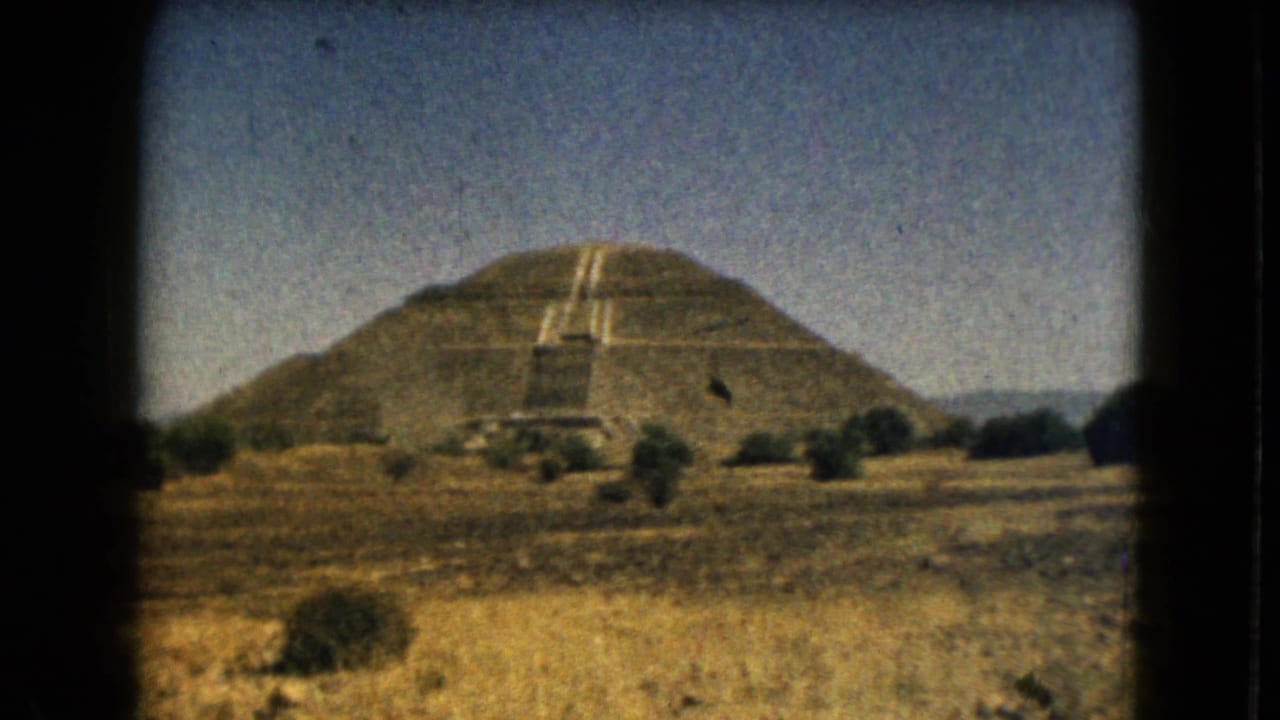 Distant stepped pyramid with central stairway, shrubs, dry field, and blue sky.