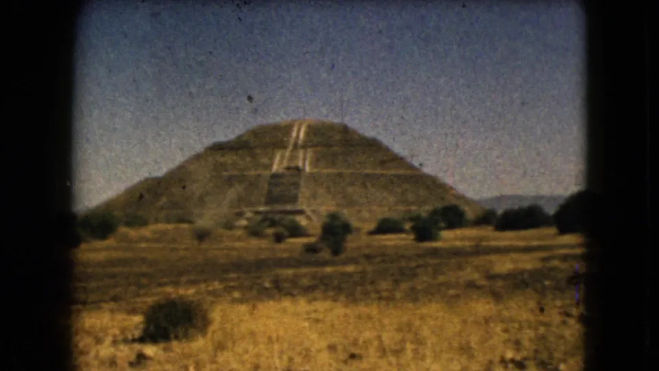 Color 8mm frame showing the pyramid slightly larger in view, with its broad stairway, dry foreground grasses, shrubs, and low hills visible.