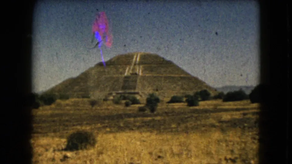 1962 8mm frame of the pyramid in an arid setting, with dry grass, shrubs, blue sky, film borders, and visible color marks from the scan.