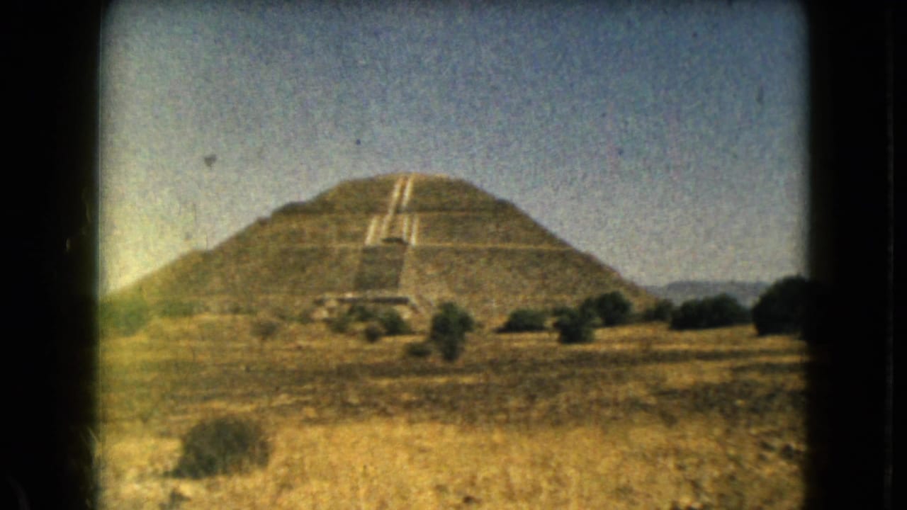 Large stepped pyramid with central staircase beyond dry grass and shrubs.