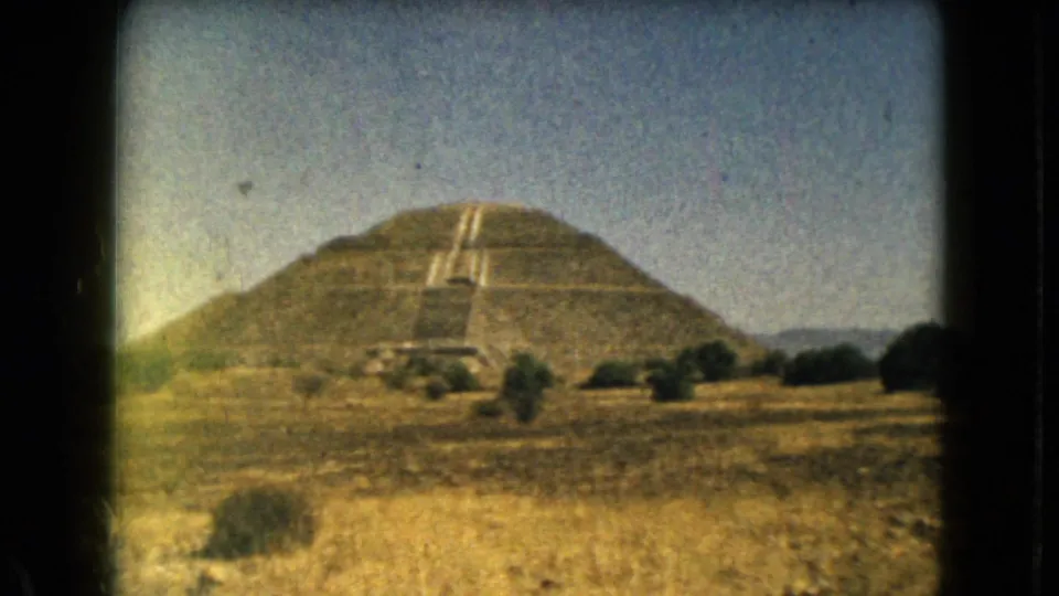 1962 8mm view near Mexico City showing a large stepped pyramid across dry grass, shrubs, and low hills under a blue sky.