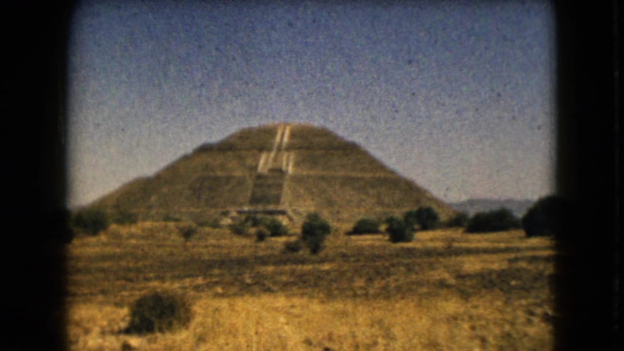 Vintage 8mm frame of a broad stepped pyramid, likely in the Mexico City area, seen from dry open terrain with sparse shrubs.