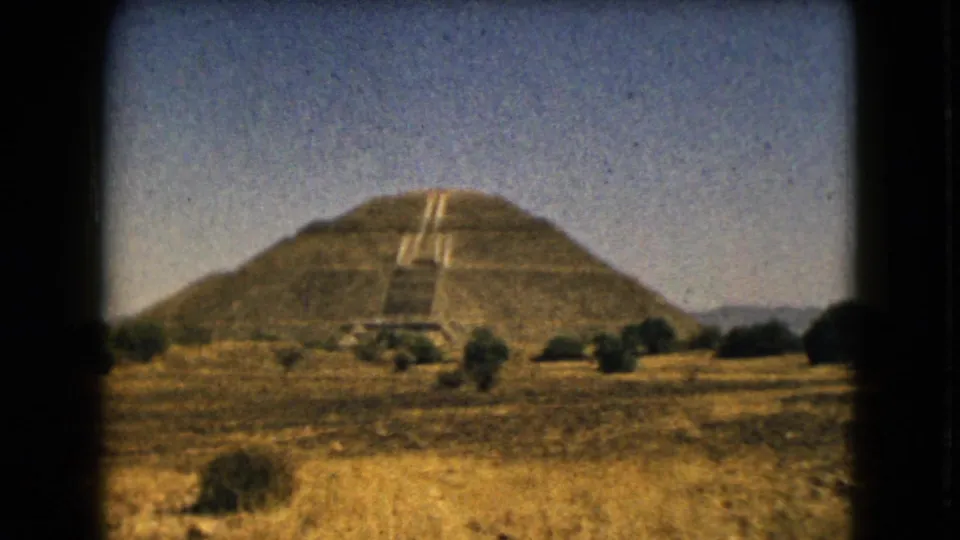 Vintage 8mm frame of a broad stepped pyramid, likely in the Mexico City area, seen from dry open terrain with sparse shrubs.