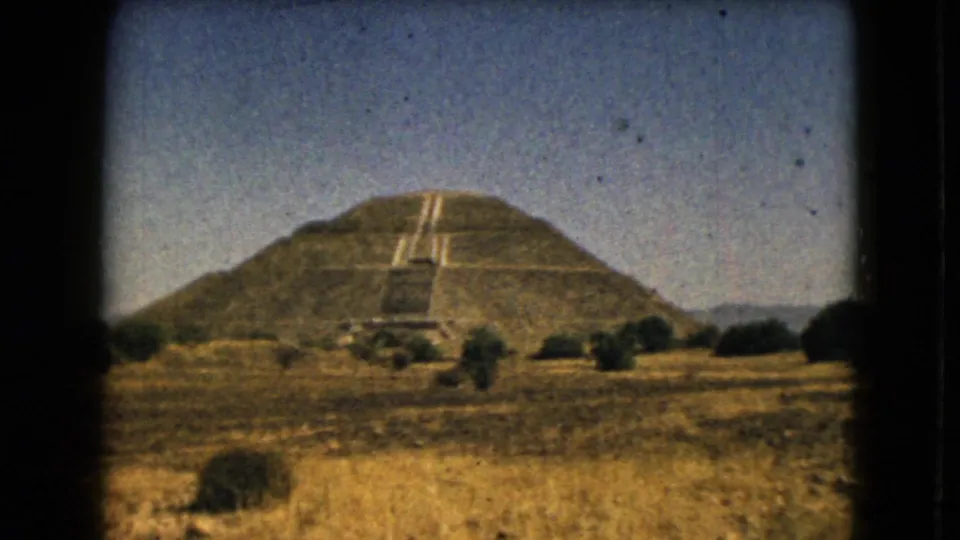 Soft 8mm frame showing the stepped pyramid from a visitor viewpoint, with yellow grass, shrubs, and a clear sky around it.