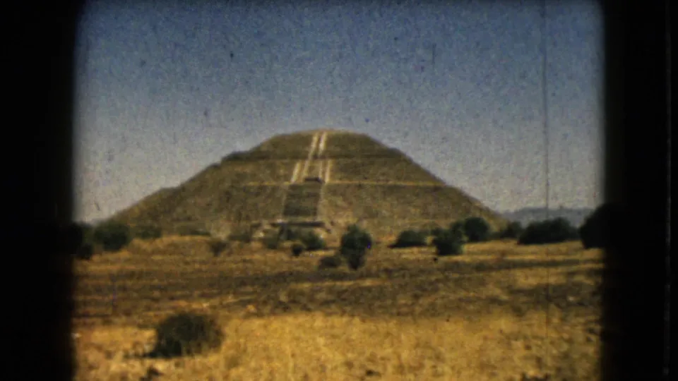 1962 home-movie frame of a stepped pyramid near Mexico City, with central stairs, dry terrain, shrubs, and visible 8mm film borders.