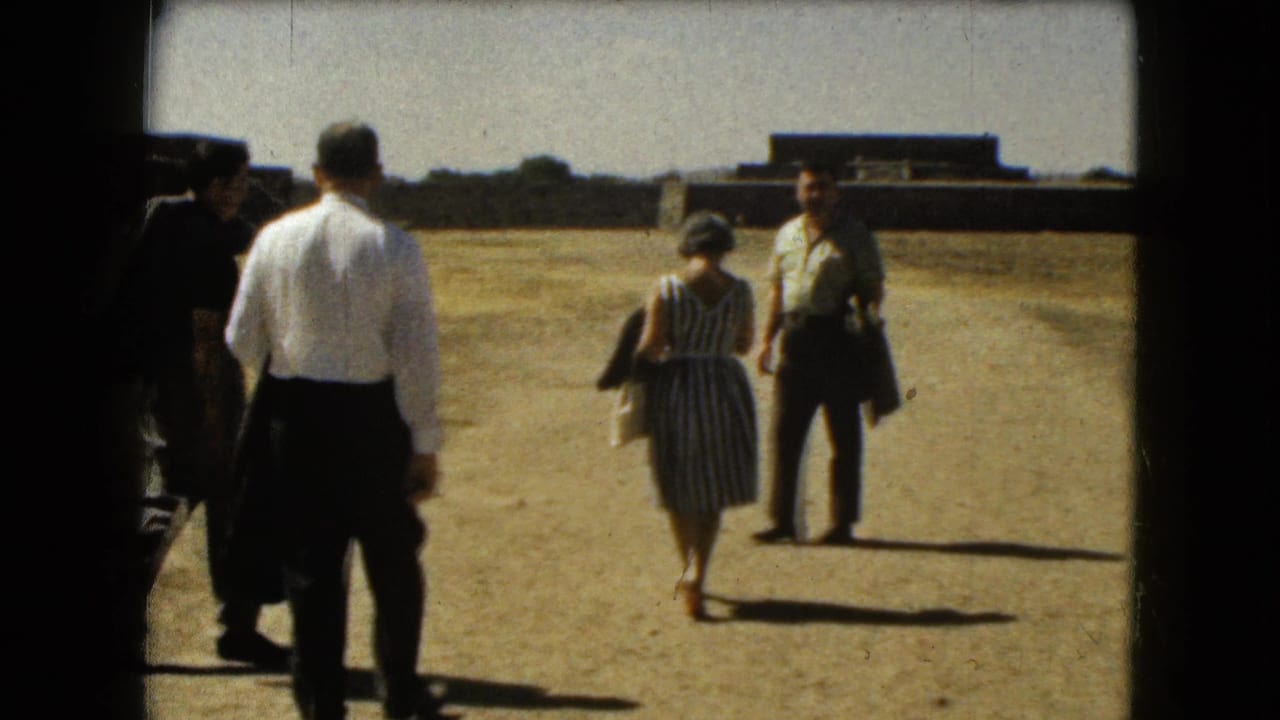 Tourists walking across a dusty field with low buildings in the distance.