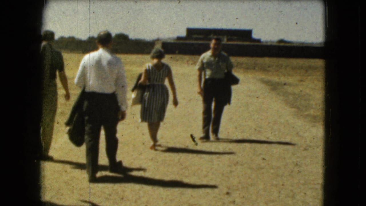 Several tourists walk on a sunlit dirt path with buildings beyond them.
