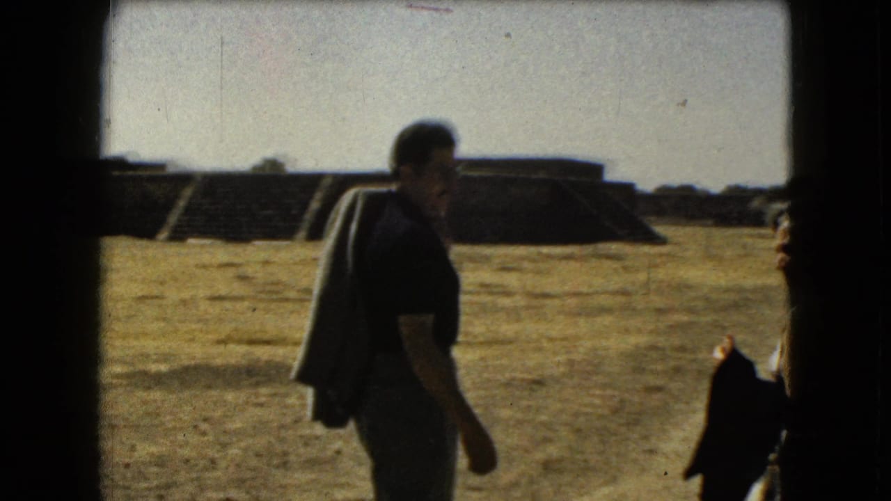 A tourist in a dark shirt walks across dry ground with stepped stone structures in the background.