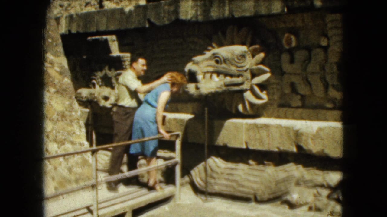 Woman in a blue dress leans toward a carved serpent-head sculpture as a man stands behind her.