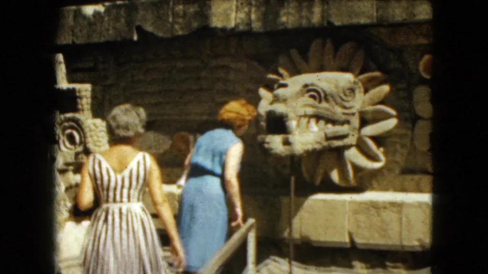 Two adult women, one in a striped dress and one in blue, view the carved serpent-head sculpture in 1962 Mexico City footage.