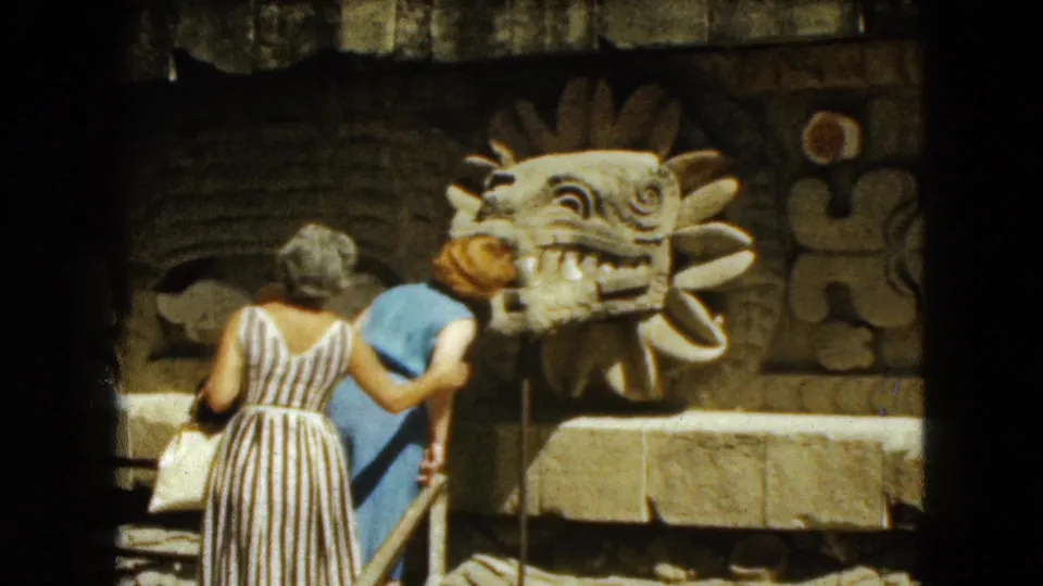 Two women stand close to the carved serpent-head sculpture and stone relief wall during a 1962 Mexico City ruins visit.