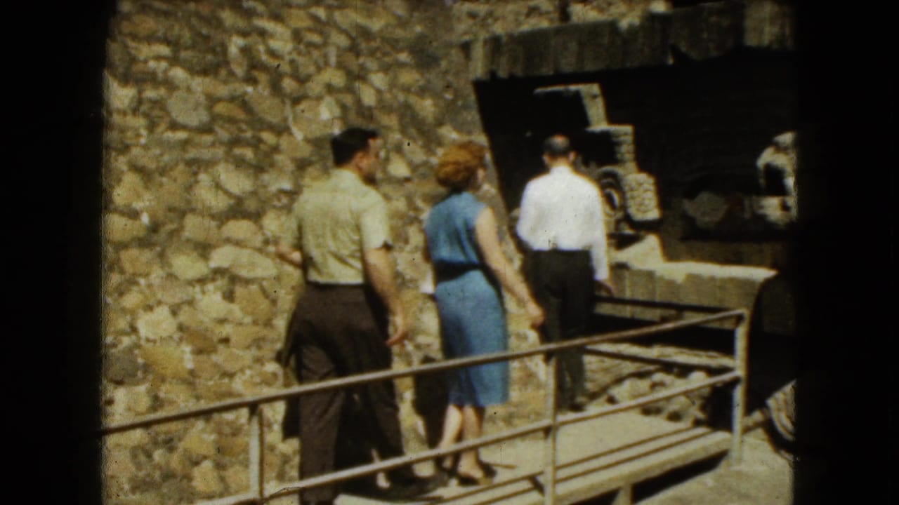 Three adults walk along a railed stone pathway beside a ruin wall.