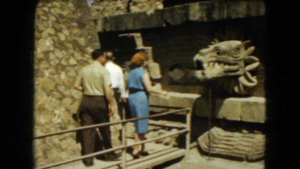 1962 Mexico City home-movie frame with adult visitors paused near a prominent carved serpent-like head at a stone ruin.
