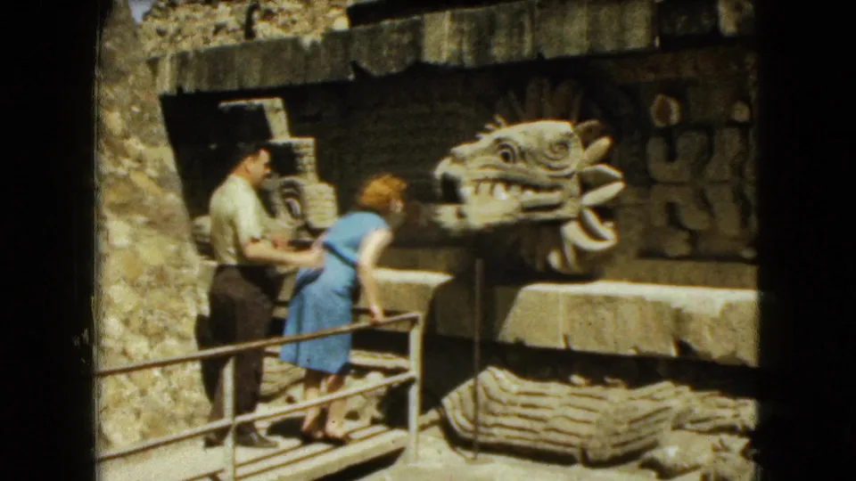 1962 Mexico City ruins footage shows a woman in a blue dress leaning over the railing while a man stands nearby by a carved stone head.