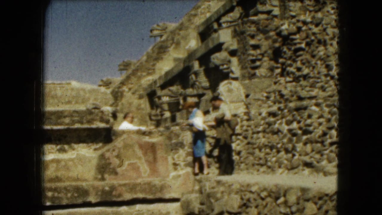 A couple stands near carved stone ruins beside a rough masonry wall.