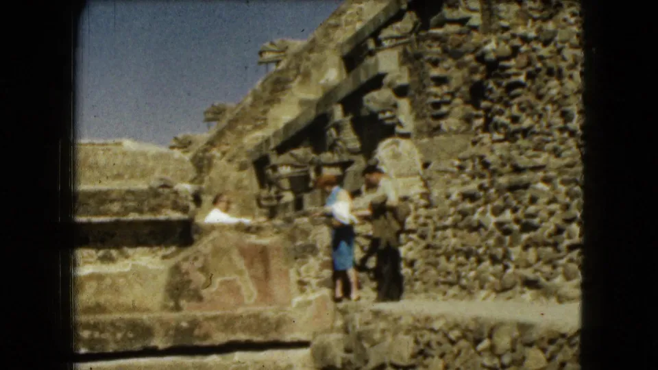 In 1962 Mexico City home-movie footage, a couple pauses beside ancient-looking carved stonework and rough masonry under a blue sky.