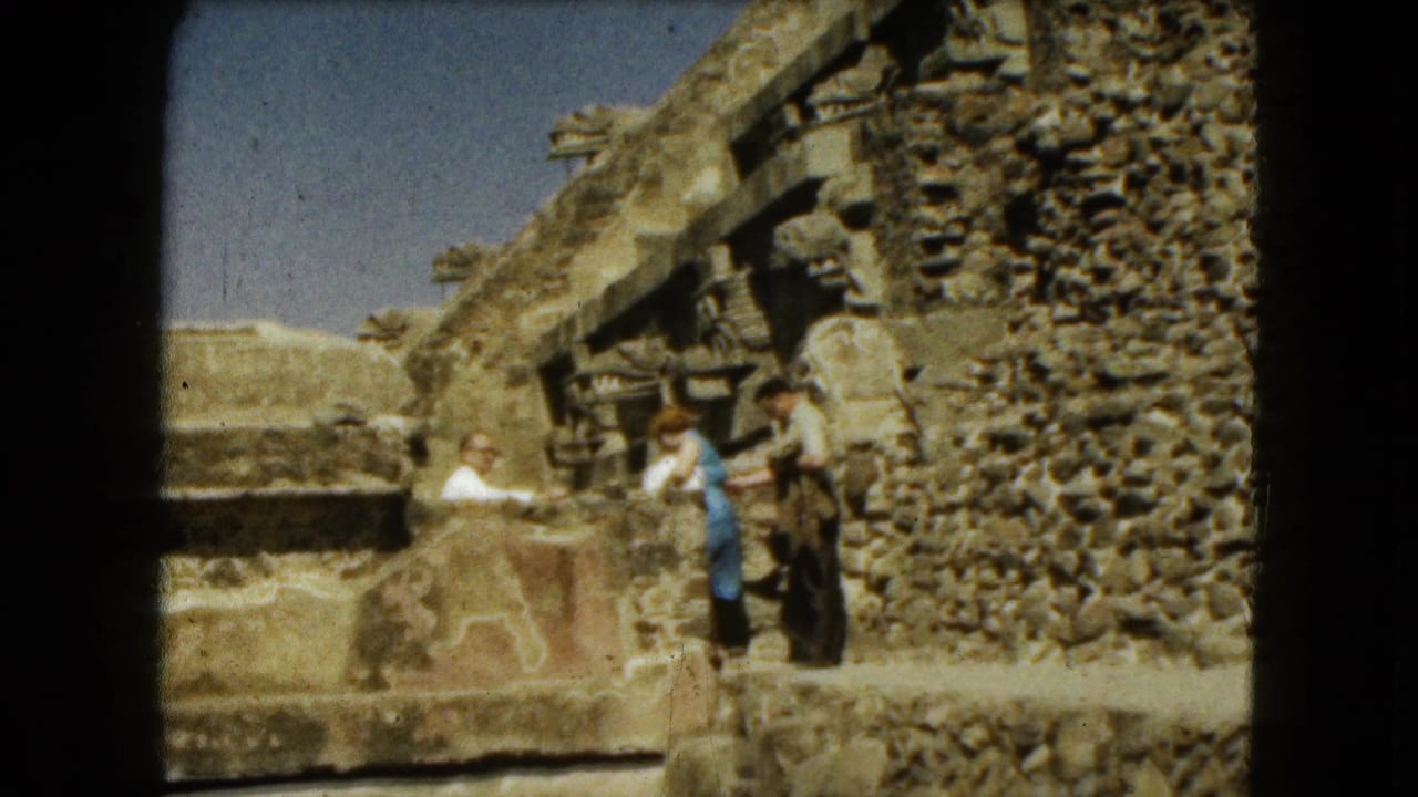 The man and woman move carefully through Mexico City ruins, framed by rough stone walls and carved architectural fragments.