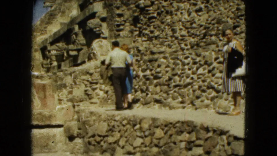 1962 Mexico City frame showing adult tourists in period clothing walking beside sunlit stone ruins.