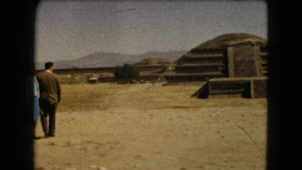 Adult visitors walk toward stepped stone ruins across a dry open archaeological site.