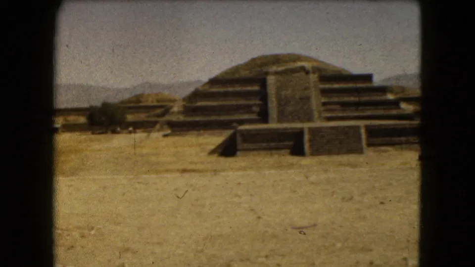 Wide 1962 Mexico City home-movie view of stepped ruins, low stone platforms, dry ground, and distant hills.