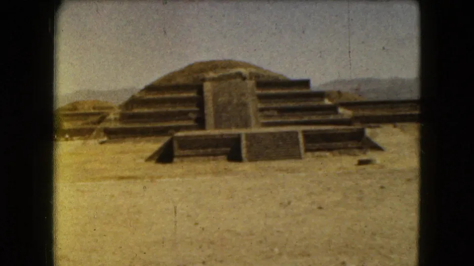 1962 8mm footage shows stepped archaeological structures in Mexico City with arid foreground and hazy hills beyond.