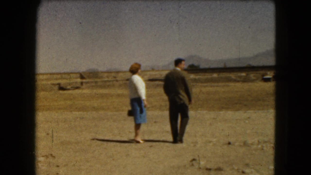 Man and woman standing on dusty ground with mountains and low ruins in the distance.