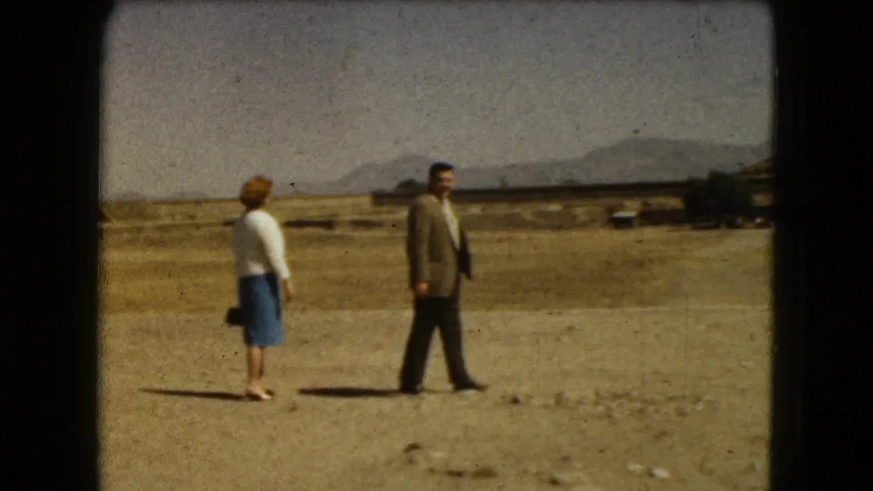 1962 8mm frame near Mexico City with a woman standing in profile as a suited man walks across an arid archaeological landscape.