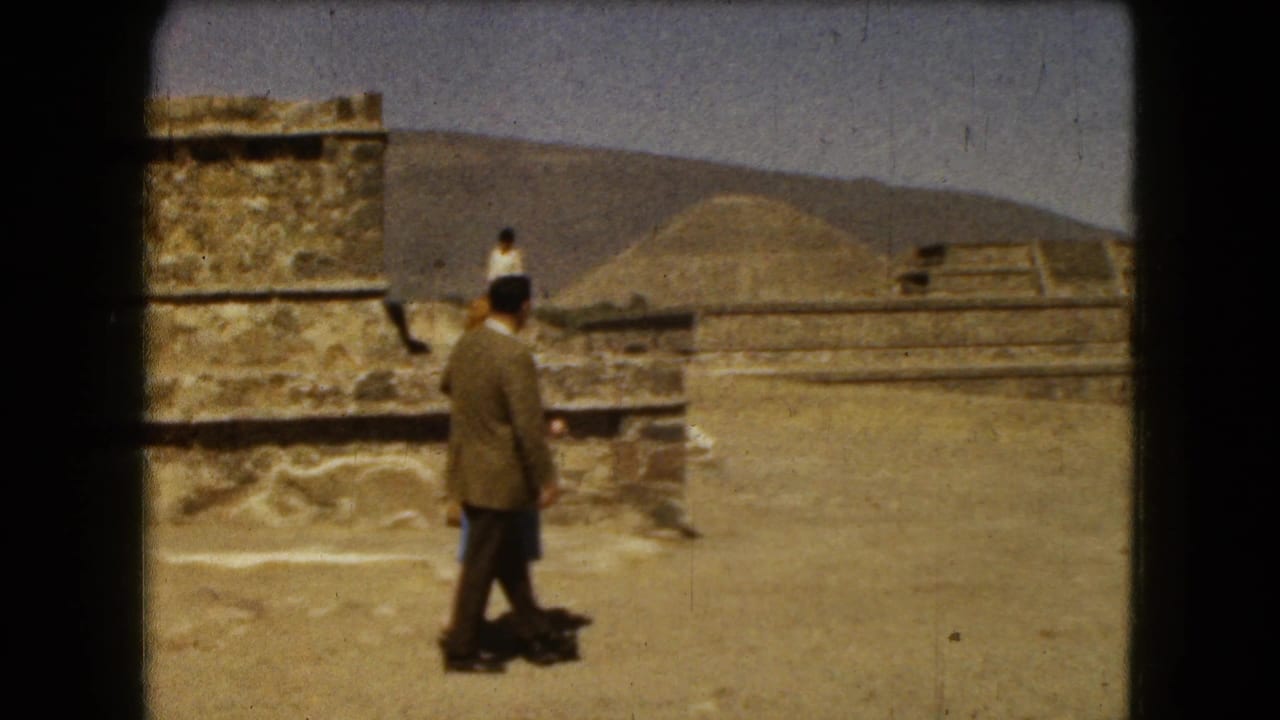 Man in a brown jacket walks across stone ruins with stepped pyramids and hills behind him.