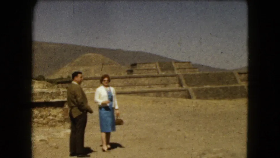 A man and woman face the 8mm camera at a Mexico City pyramid complex, with stone terraces and hills behind them.