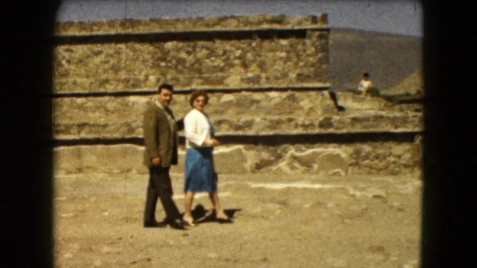 In Mexico City area travel footage from 1962, an adult couple stands near stepped stone ruins on sandy ground under a bright sky.