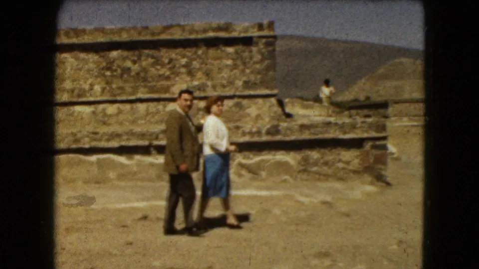 A man in a brown jacket and a woman in a white blouse and blue skirt walk past stone ruins in 1962 Mexico City travel footage.