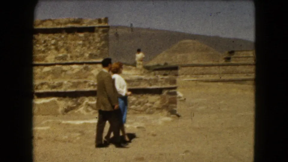 The adult couple continues across sandy ground near weathered stepped stonework, with a distant visitor visible on the ruins.