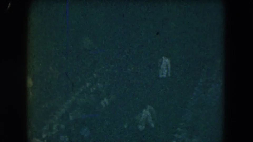 1962 Mexico City 8mm frame showing faint people or room shapes in a very dark interior with a small bright object at right.