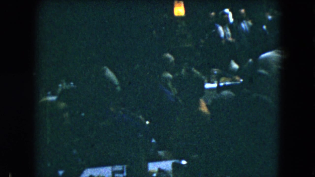 Mexico City 1962 home-movie frame of guests gathered around a table in a dark indoor room with a small warm light above.