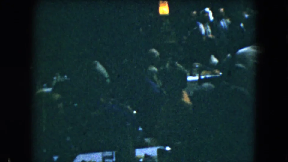 Mexico City 1962 home-movie frame of guests gathered around a table in a dark indoor room with a small warm light above.