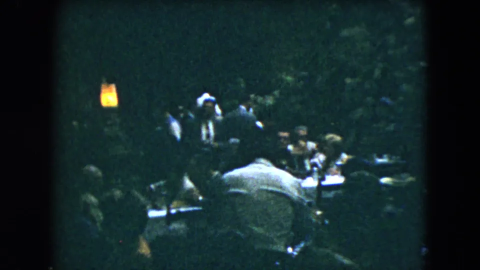 Mexico City 1962 home-movie frame with guests seated and standing in a dim crowded room, seen in soft grainy 8mm texture.