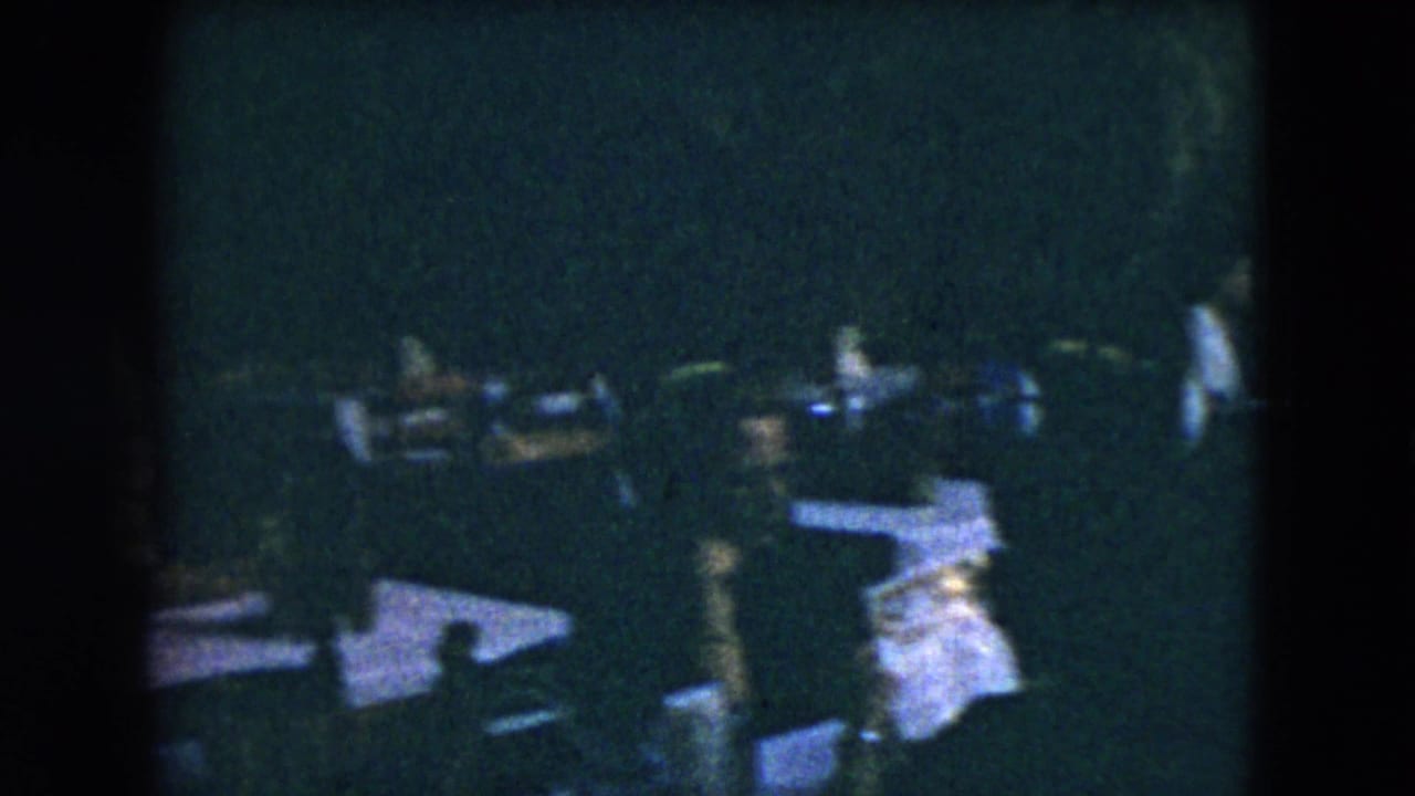 Dim outdoor dining area with white tablecloths and formally dressed guests moving around tables.