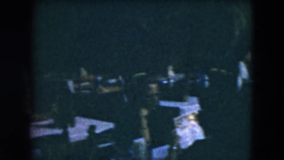 1962 Mexico City home-movie frame showing guests in formal clothing near white-clothed tables in a dim outdoor dining area.