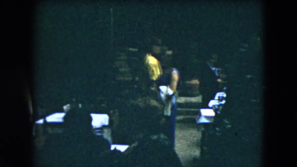 1962 Mexico City home-movie still with guests in dresses or suits standing among chairs and white-covered tables in low light.