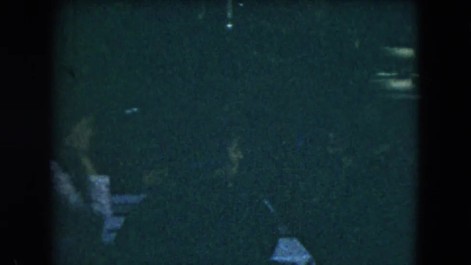 Grainy 1962 Mexico City frame showing adults seated in conversation at a small table inside a dimly lit gathering space.