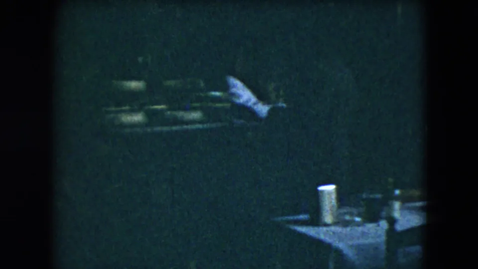 1962 Mexico City home-movie frame with a table set with cups or shakers and an adult seated nearby in a dim social room.
