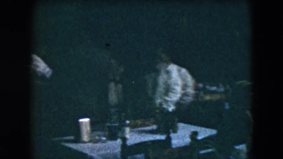Dim 1962 Mexico City frame showing adults around small tables, including a man in a light-colored suit, with tableware in front.