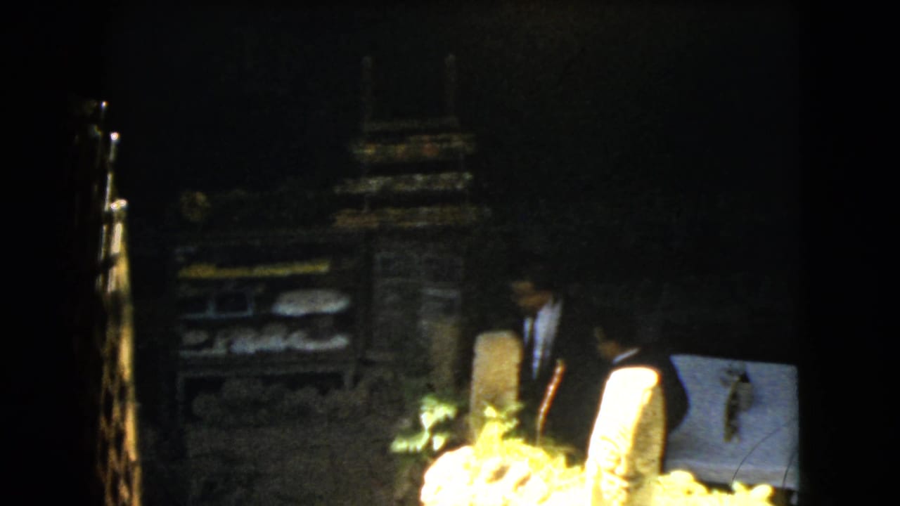 In 1962 Mexico City home-movie footage, a man in a dark suit stands near rough stonework, foliage, and a pale covered display or marker.
