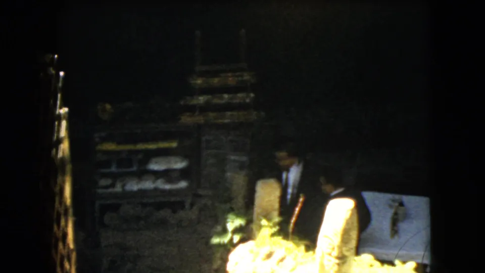 In 1962 Mexico City home-movie footage, a man in a dark suit stands near rough stonework, foliage, and a pale covered display or marker.